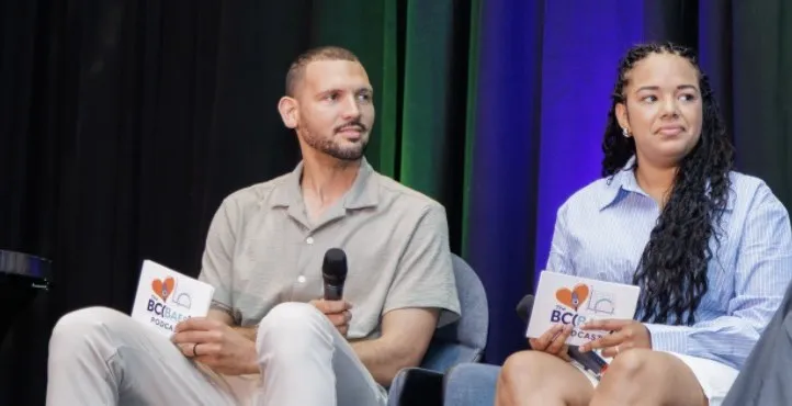 Jordan and Melina speaking on stage at BMBA Summit 2026, holding BC(BAes) Podcast cards